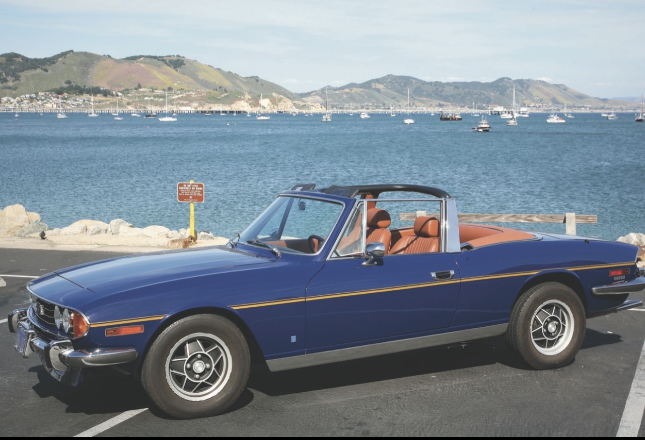 A blue Federal-specification Triumph Stag convertible, showing the wing side marker lamps and the door mirror.