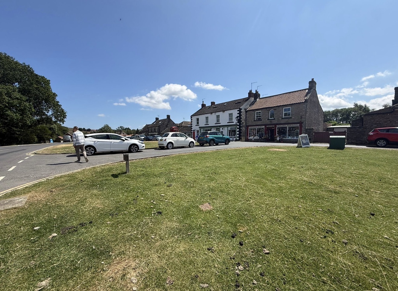 The wide grass village green at Goathland with a stone terrace of shops beyond and a red telephone box at the roadside.