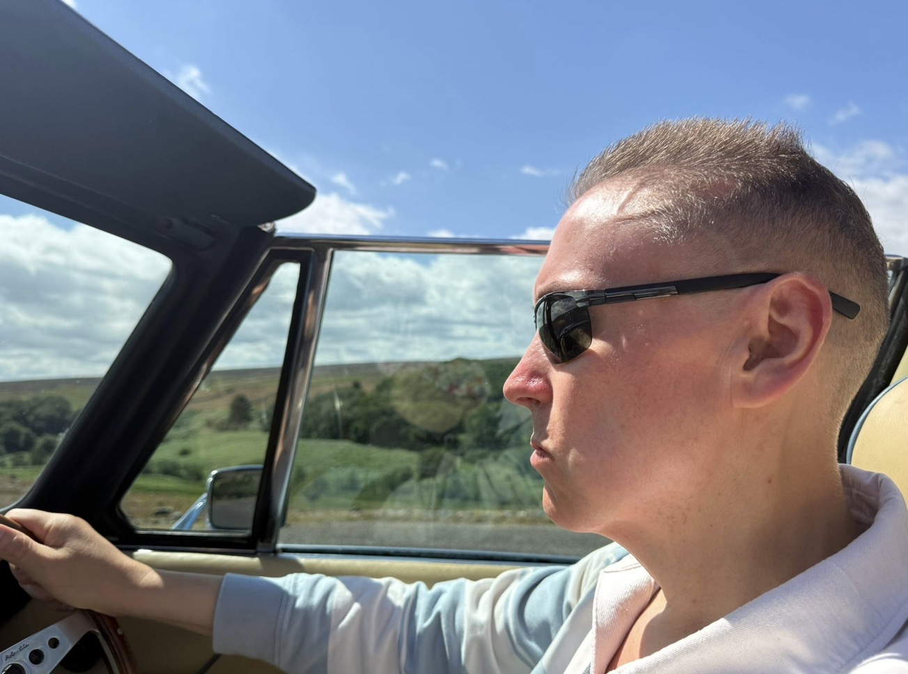 The driver of a Triumph Stag at the wheel, the roof down, moorland and green pasture open to the side.