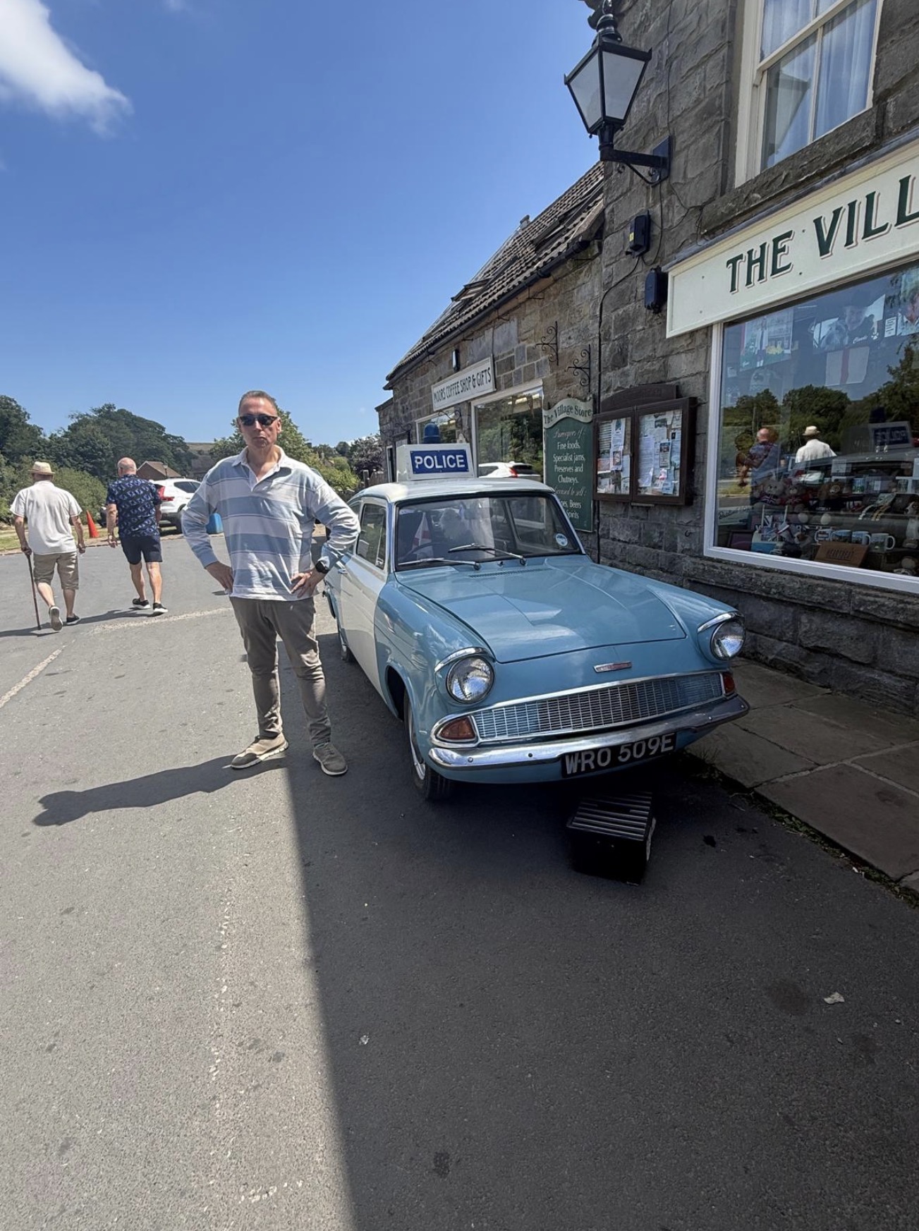 A pale blue and white Ford Anglia police car, registration WRO 509E, with a POLICE sign on the roof, parked on the pavement outside the Goathland village store.